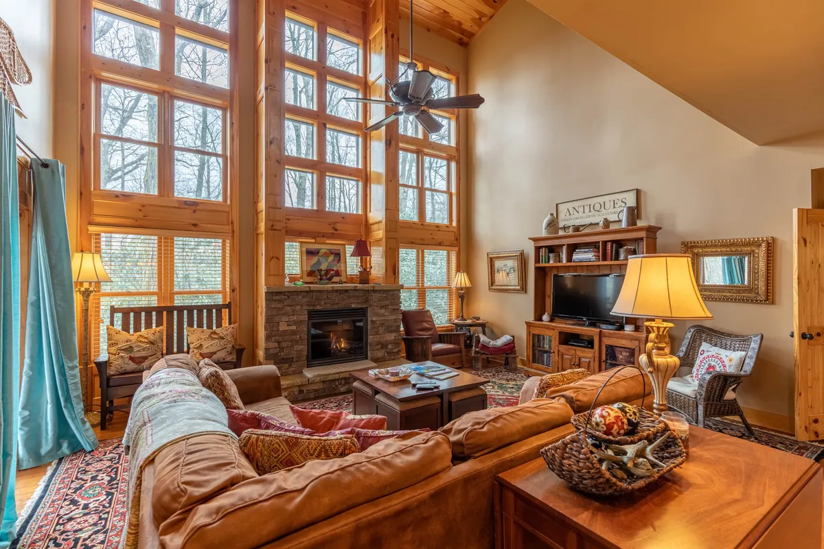 This image showcases a spacious living room with soaring vaulted ceilings, large windows that let in abundant natural light, a cozy fireplace, comfortable suede seating, exposed wooden beams, and tasteful decorative accents. The view through the windows reveals lush greenery, likely rhododendrons, enhancing the connection to the outdoors.