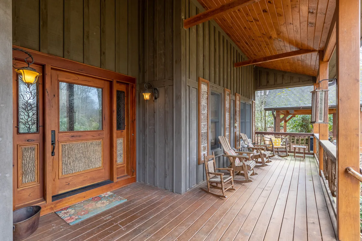This image shows a welcoming front porch with wooden rocking chairs, a sturdy wooden railing, and decorative light fixtures. The porch faces out toward nature, with gentle sunlight illuminating the space and the wooden front door adding warmth to the scene.