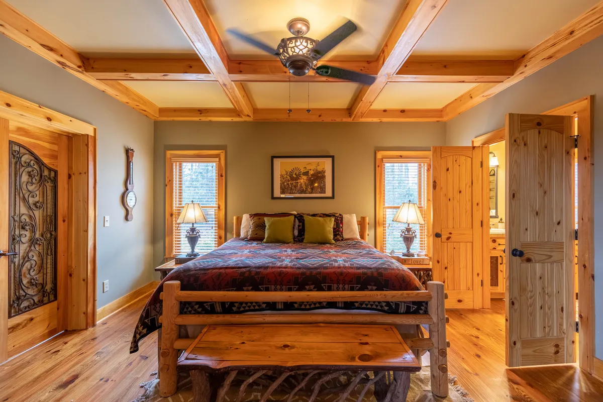 A warmly lit bedroom featuring a bed with patterned bedding, a wooden bench at the foot, a window letting in natural light, and tasteful wall art. The overall mood is calm and restful, perfect for quiet relaxation after a busy day.