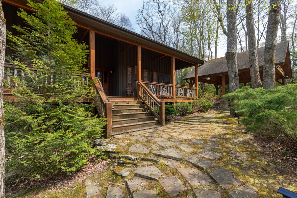 A tranquil exterior scene showcasing a wooden porch, stone pathway, and lush greenery surrounding the home. Broad steps lead up to the inviting entrance, framed by natural landscaping ideal for relaxed outdoor moments.