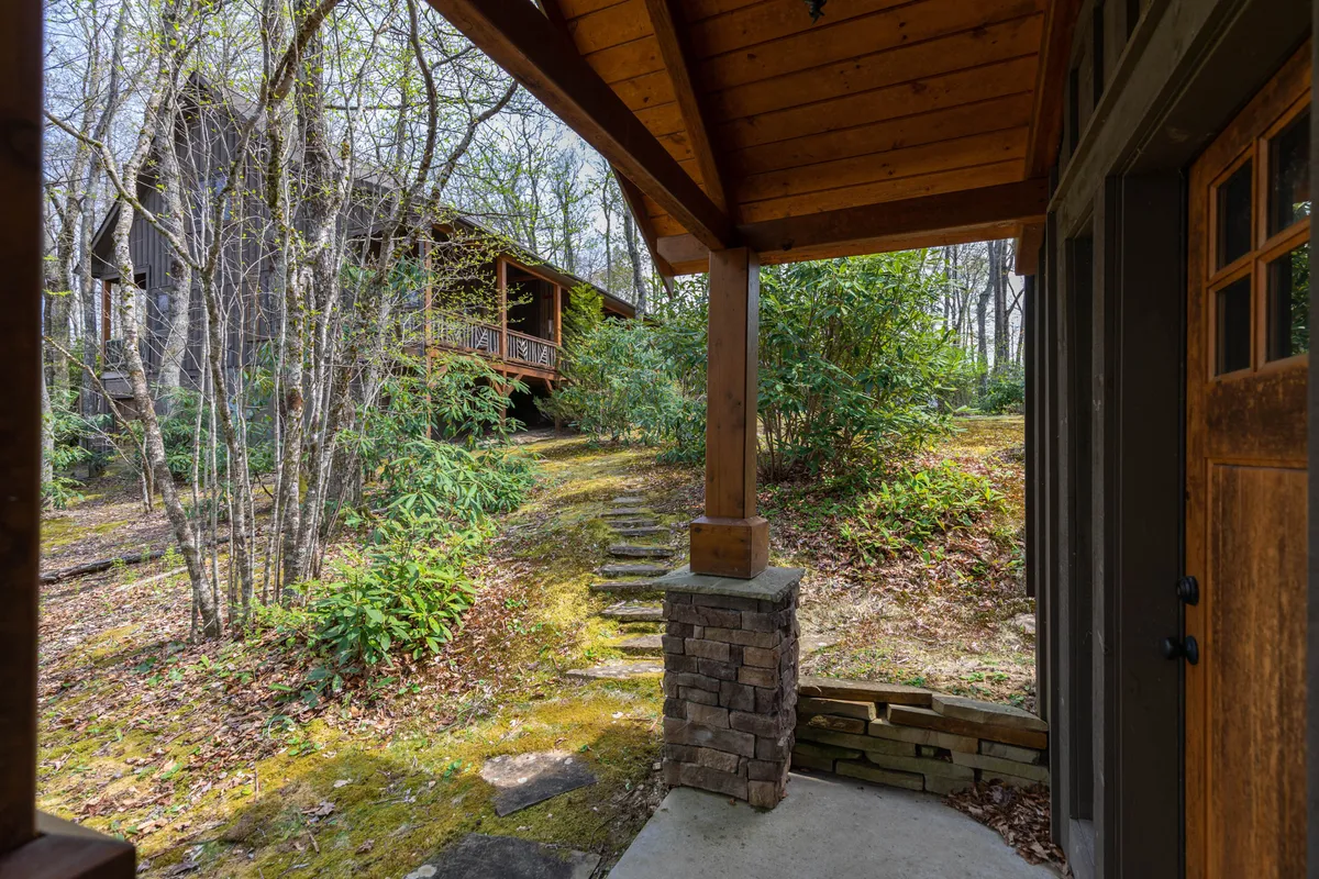 A peaceful wooden porch with stone pillar, bordered by steps leading directly into the woods. The scene is enveloped in greenery and sunlight, evoking a sense of calm and exploration from the doorstep.