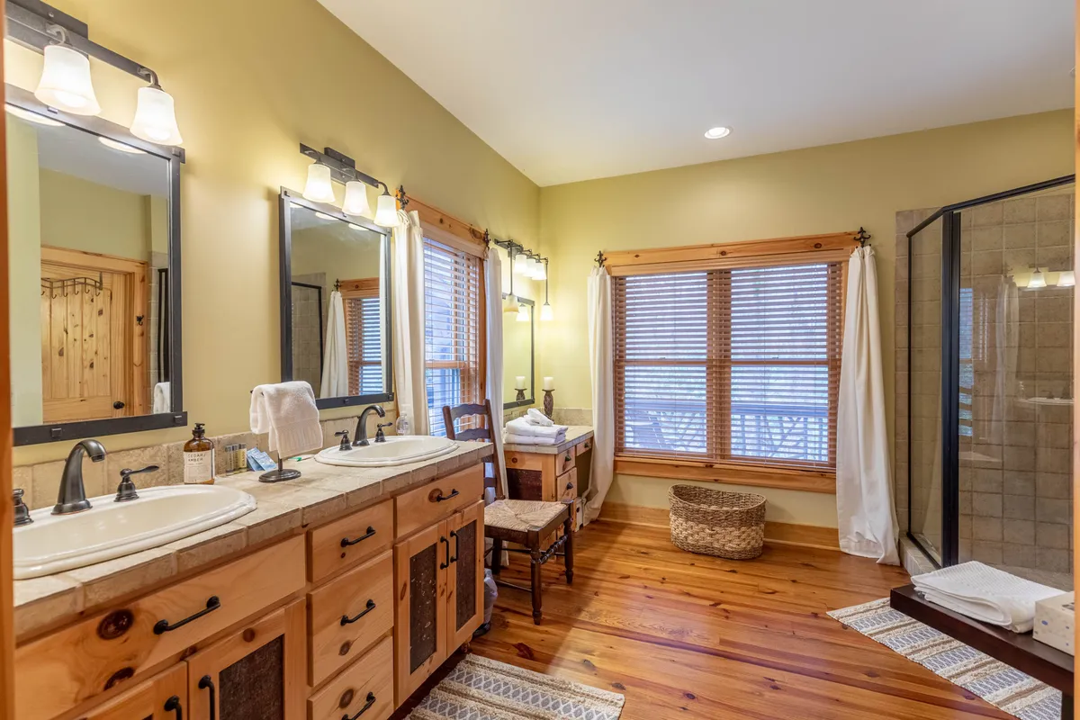 A serene bathroom featuring double vanity sinks, large windows, natural wood cabinetry, and a spacious shower area. The design is calm and spa-like, emphasizing privacy and convenience.