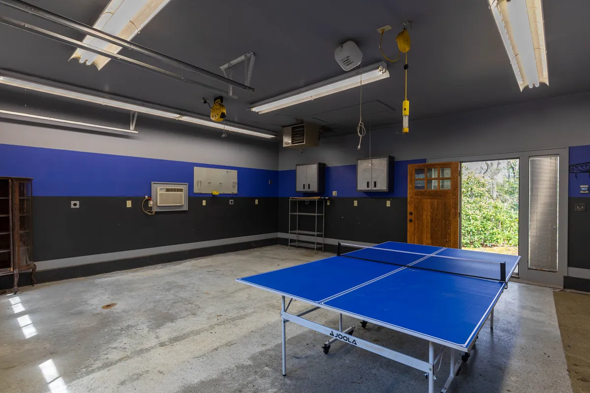 This image showcases the recreation bay of the garage, featuring a prominent ping pong table set against blue and grey walls, with a wooden door visible in the background. The space is set up for energetic activity, with enough room for friendly games or casual gatherings. Some gear and equipment are visible, hinting at storage options for outdoor adventures.