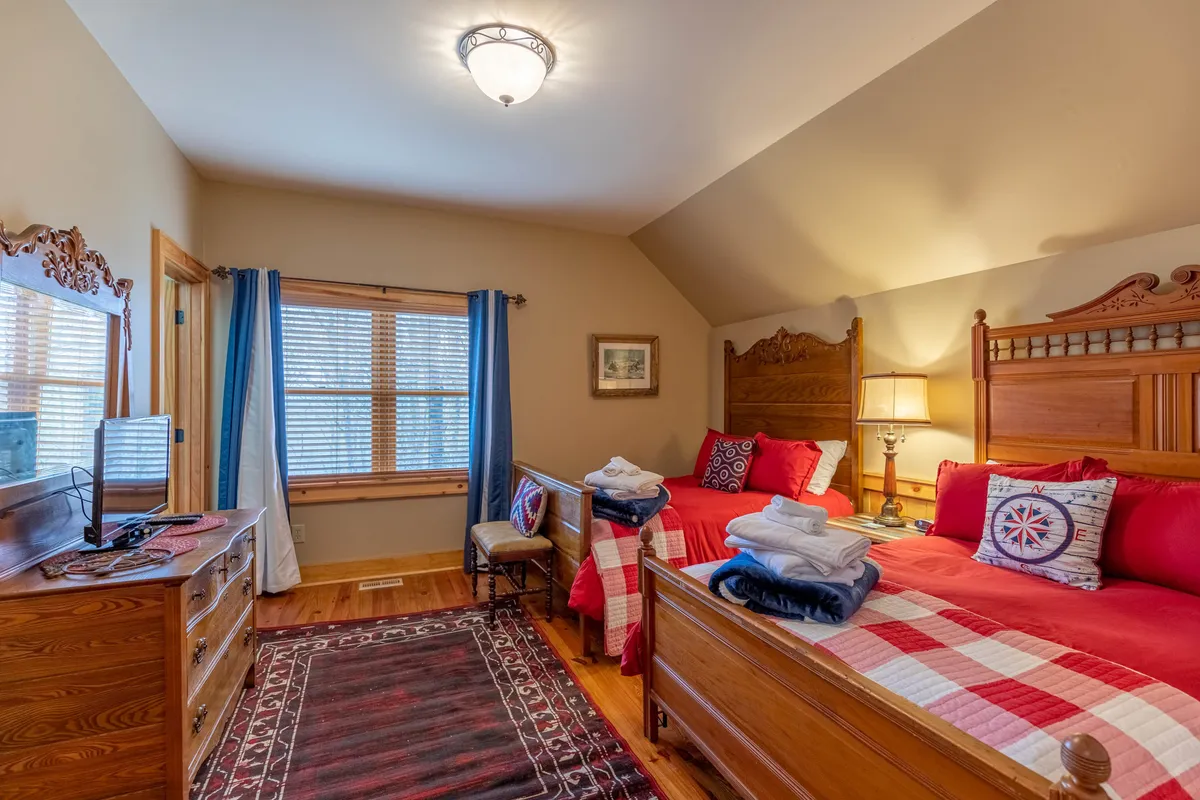 A comfortable, sunlit bedroom with twin beds decorated with colorful pillows, a large window with curtains, a wooden dresser, and a cozy area rug. The ambiance is cheerful and inviting, making it ideal for teens or friends.