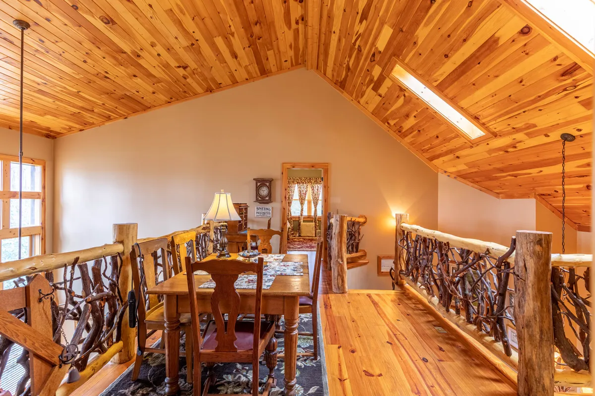 A dining area centered around a substantial wooden table, with rustic railings, a skylight above, and a cozy lamp. The setting is designed for family meals, board games, and storytelling, with a warm, inviting ambiance ideal for group gatherings.