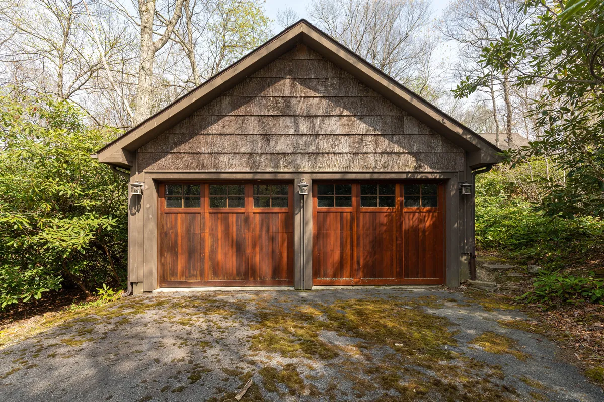 An exterior view of the garage’s wooden doors surrounded by greenery and a shingle roof. The tranquil setting hints at the garage’s location down a mossy path and its connection to the outdoors. The image suggests a practical entry point for both vehicles and gear.