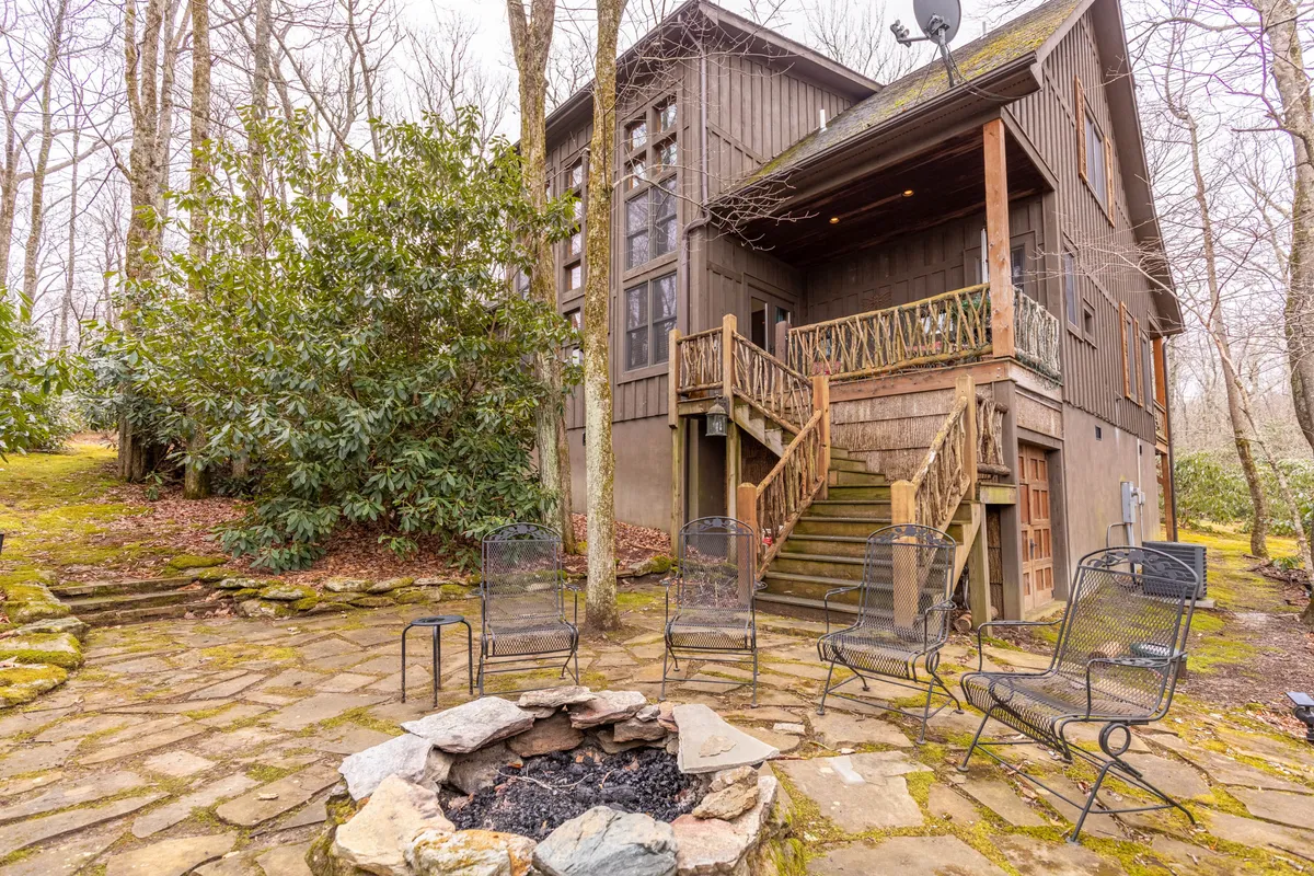 A stone fire pit is the focal point, encircled by a seating area and bordered by tall trees. Wooden stairs lead from the deck area to this lower gathering spot. The ambiance is cozy and communal, designed for storytelling, s’mores, and time under the stars.
