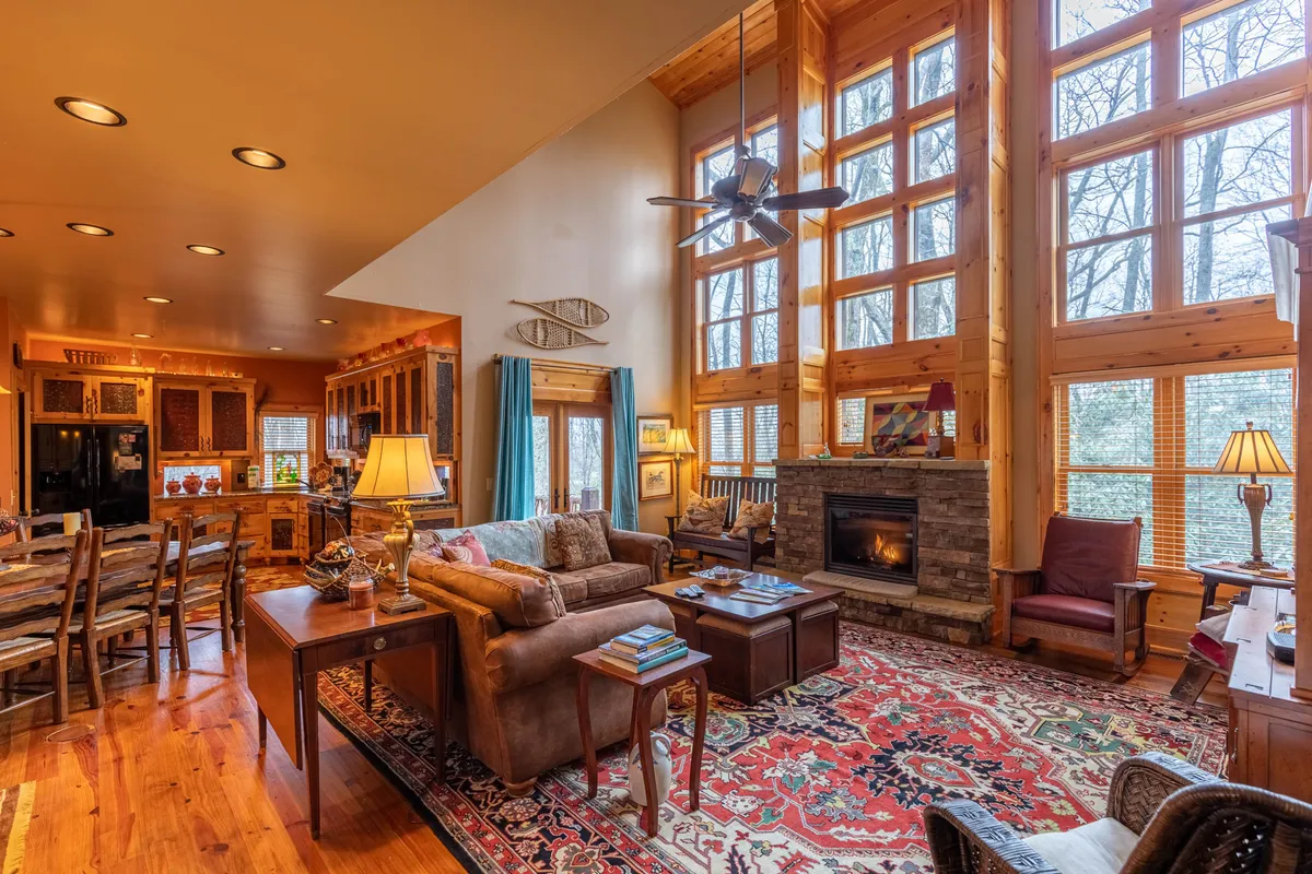 A warm, open-plan space featuring a large windowed living area with a fireplace, a soft rug, a comfy sofa, and a view into the adjacent kitchen area. Sunlight streams through the windows, emphasizing the home’s airy, connected layout and fostering a sense of togetherness.
